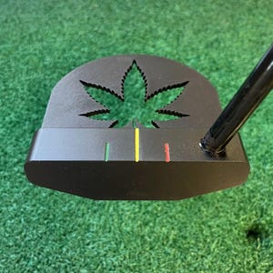 Könnte beinhalten: Schwarzer Golfschläger mit einem Cannabisblatt-Ausschnitt. Der Putter hat einen schwarzen Schaft und einen Kopf mit grünen, gelben und roten Ausrichtungslinien. Der Putter befindet sich vor einem grünen Rasenhintergrund.