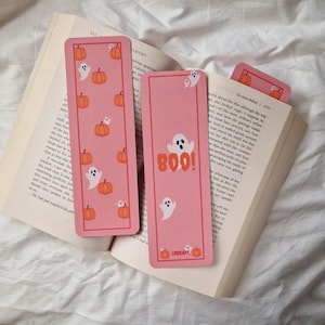 Op de afbeelding: Roze Halloween-thema bladwijzers met pompoen- en spookillustraties. Eén bladwijzer heeft het woord "BOO!" in oranje letters. De bladwijzers liggen op een open boek.
