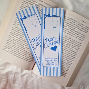 Könnte beinhalten: Zwei blau-weiß gestreifte Lesezeichen mit dem Text "Team Conrad" und einem blauen Herz. Ein Lesezeichen hat das Zitat "when the sun comes out, all the stars disappear". Die Lesezeichen liegen auf einem offenen Buch.
