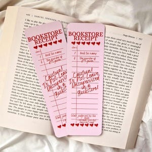 Op de afbeelding: Twee roze "Bookstore Receipt" bladwijzers met rode hartaccenten en handgeschreven tekst, geplaatst op een open boek. De bladwijzers luiden "Let op! Niet onbeheerd achterlaten in een boekhandel!"