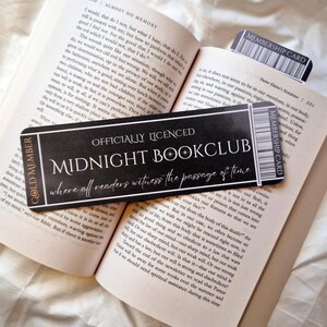 May include: A black and gold bookmark with the text "Officially Licensed Midnight Bookclub where all readers witness the passage of time". The bookmark is on top of an open book with text about good and evil.