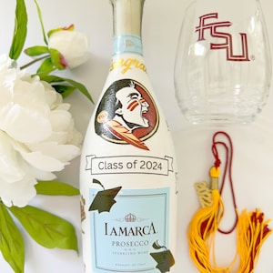 May include: A white bottle of LaMarca Prosecco sparkling wine with a graduation cap and FSU Seminole logo design. The bottle has a gold tassel and the text "Class of 2024" and "Noles".