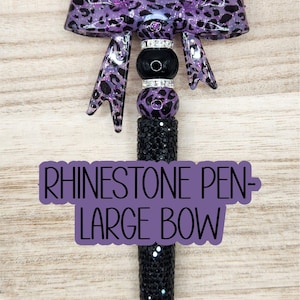May include: A rhinestone pen with a large bow. The pen features a black, glittery barrel and a decorative top with a purple leopard print bow. The pen also has black and silver beads.