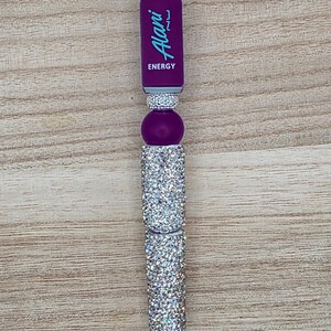 K&ouml;nnte beinhalten: Ein Kugelschreiber mit einem violetten Schaftabschnitt und dem Text Alani Nu ENERGY in Hellblau. Der Stift ist mit mehreren silberfarbenen, mit Strasssteinen besetzten Segmenten und einer violetten, kugelf&ouml;rmigen Perle im oberen Bereich verziert.
