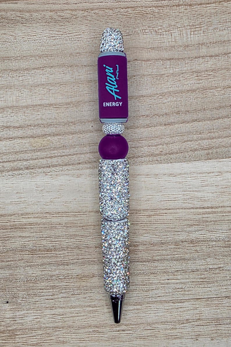 K&ouml;nnte beinhalten: Ein Kugelschreiber mit einem violetten Schaftabschnitt und dem Text Alani Nu ENERGY in Hellblau. Der Stift ist mit mehreren silberfarbenen, mit Strasssteinen besetzten Segmenten und einer violetten, kugelf&ouml;rmigen Perle im oberen Bereich verziert.