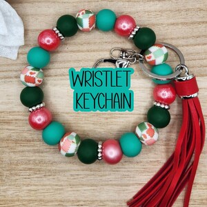 May include: A circular wristlet keychain featuring alternating matte and metallic beads in shades of red, dark green, and teal, interspersed with patterned beads. A silver metal clasp and a long red faux leather tassel are attached to the ring.