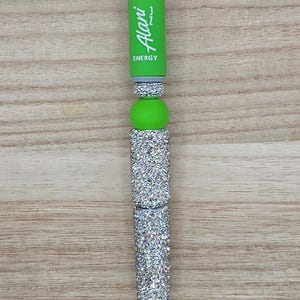 K&ouml;nnte beinhalten: Ein individueller Stift mit einem lindgr&uuml;nen Schaft, auf dem der wei&szlig;e Schriftzug Alani ENERGY vertikal aufgedruckt ist. Der Stift ist mit mehreren Abschnitten aus silberfarbenen Strasssteinen und einer glatten lindgr&uuml;nen Perle verziert.