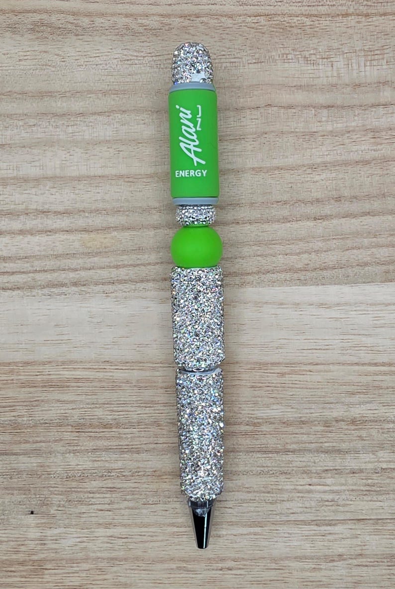 K&ouml;nnte beinhalten: Ein individueller Stift mit einem lindgr&uuml;nen Schaft, auf dem der wei&szlig;e Schriftzug Alani ENERGY vertikal aufgedruckt ist. Der Stift ist mit mehreren Abschnitten aus silberfarbenen Strasssteinen und einer glatten lindgr&uuml;nen Perle verziert.