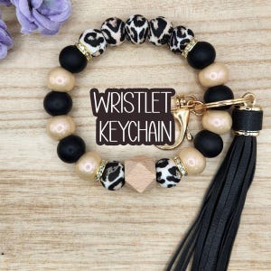 May include: Wristlet keychain with a black leather tassel. The beaded bracelet features black, tan, and leopard print beads, with gold accents and a wooden cube. The text "Wristlet Keychain" is displayed.