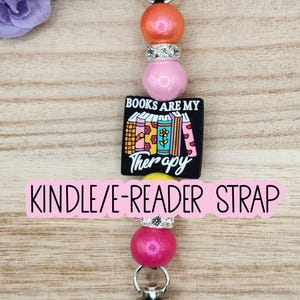 May include: A Kindle/e-reader strap featuring a silver metal lobster clasp at each end, strung with colorful beads including orange, pink, and yellow spheres, two rhinestone-encrusted spacers, and a black square charm with books and text.