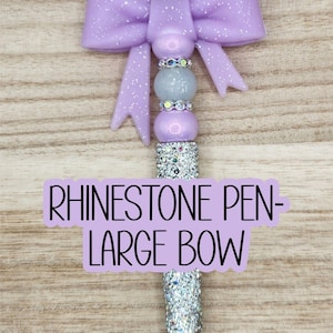 May include: A decorative pen with a large, glittery purple bow and a silver rhinestone barrel. The pen has a black tip and the text "RHINESTONE PEN - LARGE BOW" on a purple label.