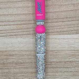 K&ouml;nnte beinhalten: Ein einziehbarer Stift mit einem rosa Schaft, auf dem der Text Alani Nu ENERGY in Hellblau gedruckt ist. Der Stiftk&ouml;rper ist mit abwechselnden Abschnitten aus silberfarbenen Strasssteinen und glatten rosa Perlen verziert und hat eine silberne Metallspitze.