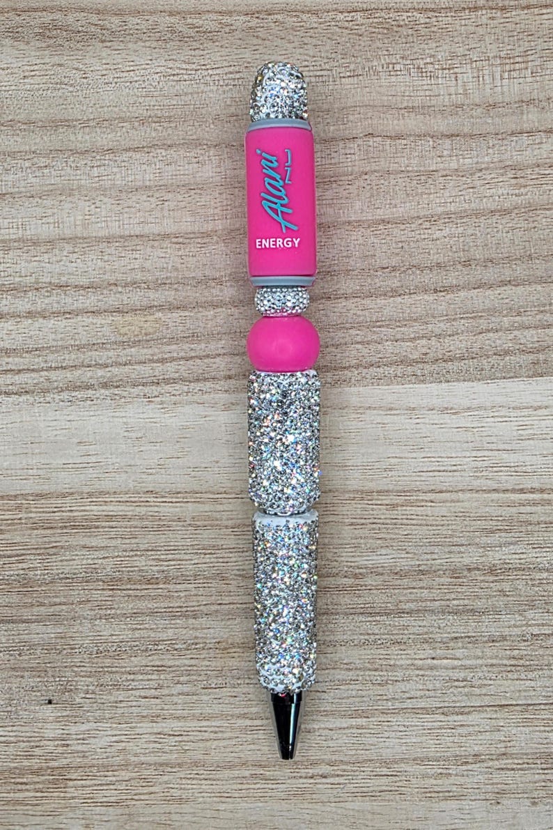 K&ouml;nnte beinhalten: Ein einziehbarer Stift mit einem rosa Schaft, auf dem der Text Alani Nu ENERGY in Hellblau gedruckt ist. Der Stiftk&ouml;rper ist mit abwechselnden Abschnitten aus silberfarbenen Strasssteinen und glatten rosa Perlen verziert und hat eine silberne Metallspitze.