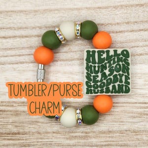 May include: A circular charm featuring olive green and orange silicone beads, interspersed with silver-toned spacers encrusted with clear rhinestones. A square, olive green silicone pendant is attached, displaying the text HELLO HUNTING SEASON GOODBYE HUSBAND in a wavy, dark green font.