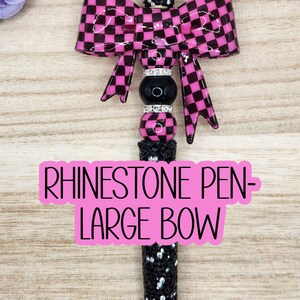 May include: A black and pink rhinestone pen with a large bow. The bow features a checkered pattern. The pen has a black barrel covered in rhinestones. The text "RHINESTONE PEN - LARGE BOW" is on the pen.