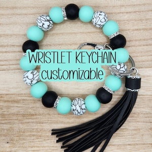 May include: A wristlet keychain with a black leather tassel. The keychain features a series of beads in black, mint green, and white with a black and white pattern. The text "WRISTLET KEYCHAIN customizable" is displayed on the keychain.