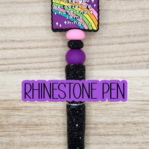 May include: A black rhinestone pen with a black tip. The pen has a decorative top with a rainbow design and the text "I like you because we hate the same things." The pen is approximately 6 inches long.