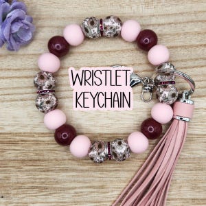 May include: A wristlet keychain with a pink tassel and a mix of pink, burgundy, and floral-patterned beads. The text "WRISTLET KEYCHAIN" is displayed on a white background. The keychain is designed to be worn on the wrist.