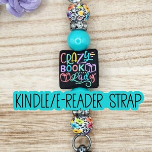 May include: A beaded strap featuring a black square charm with the text CRAZY BOOK LADY and book icons. The design includes turquoise beads, floral patterned beads, and silver-toned rhinestone spacers, finished with two dark metal lobster clasps.