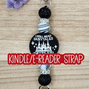 May include: A beaded strap featuring a round black charm with the white text I STILL READ FAIRYTALES and a castle graphic. The strap includes black speckled beads, white and black marbled beads, and silver-toned rhinestone spacers with metal clips.
