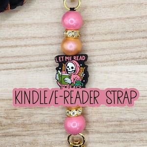 May include: A beaded Kindle or e-reader strap featuring a gold-tone metal clasp at each end. The design includes pink and gold spherical beads, two rhinestone-encrusted spacers, and a central charm depicting a skeleton reading with the text LET ME READ IN PEACE.