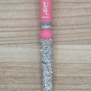K&ouml;nnte beinhalten: Ein einziehbarer Stift mit einem rosa Schaft, auf dem vertikal der wei&szlig;e Schriftzug Alani ENERGY aufgedruckt ist. Der Stift ist mit mehreren Abschnitten aus silberfarbenen Strasssteinen und einer glatten rosa Perle verziert.