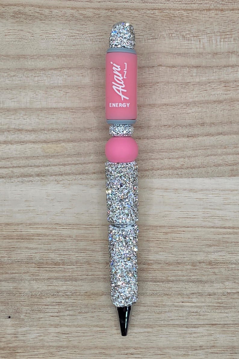K&ouml;nnte beinhalten: Ein einziehbarer Stift mit einem rosa Schaft, auf dem vertikal der wei&szlig;e Schriftzug Alani ENERGY aufgedruckt ist. Der Stift ist mit mehreren Abschnitten aus silberfarbenen Strasssteinen und einer glatten rosa Perle verziert.