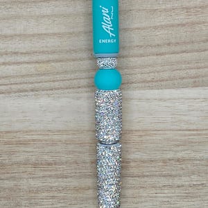 K&ouml;nnte beinhalten: Ein Kugelschreiber mit einem t&uuml;rkisfarbenen Schaft, auf dem vertikal der wei&szlig;e Schriftzug Alani ENERGY aufgedruckt ist. Der Stift ist mit mehreren silbernen, glitzernden zylindrischen Perlen und einer glatten t&uuml;rkisfarbenen Silikonperle verziert.