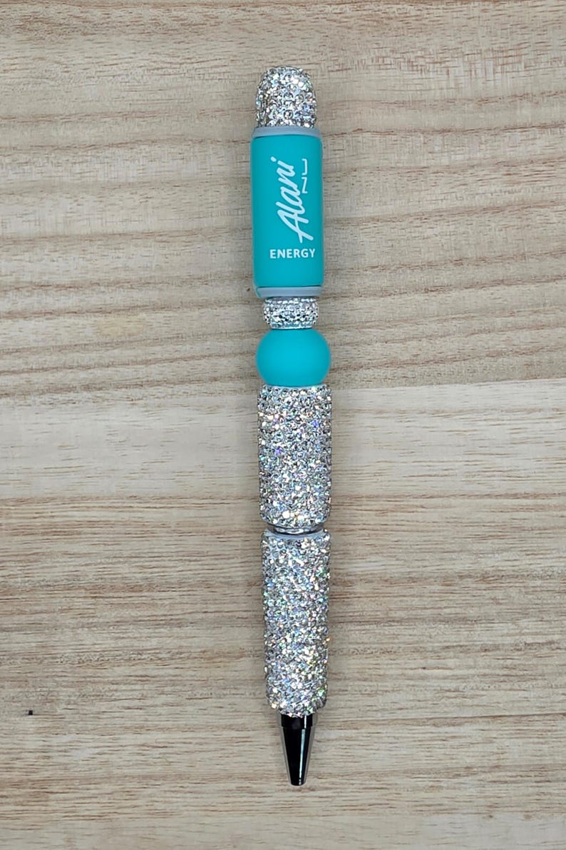 K&ouml;nnte beinhalten: Ein Kugelschreiber mit einem t&uuml;rkisfarbenen Schaft, auf dem vertikal der wei&szlig;e Schriftzug Alani ENERGY aufgedruckt ist. Der Stift ist mit mehreren silbernen, glitzernden zylindrischen Perlen und einer glatten t&uuml;rkisfarbenen Silikonperle verziert.