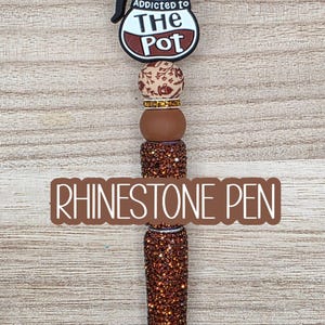 May include: A pen featuring a coffee pot topper with the text Addicted to The Pot, followed by a series of beads including a brown rhinestone sphere, a floral patterned bead, a gold spacer, a solid brown bead, and two brown rhinestone cylinders.