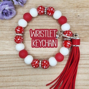 May include: A wristlet keychain with red and white beads. The red beads have a heart pattern. The keychain has a red tassel and the text "Wristlet Keychain" in a red box.