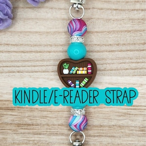 May include: A Kindle/E-Reader strap featuring a central brown heart-shaped bookshelf charm with miniature books and a plant. The strap includes two silver-toned metal clasps, two turquoise beads, two silver rhinestone spacers, and two swirl-patterned beads in shades of pink, blue, and purple.