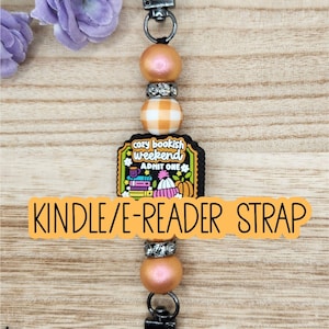 May include: A Kindle/e-reader strap featuring a central rectangular charm with the text "cozy bookish weekend ADMIT ONE" surrounded by illustrations of books, a hat, and a pumpkin. The strap includes peach-colored beads and silver-toned spacers.