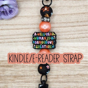 May include: A beaded strap featuring a black rectangular charm with the text A WELL READ WOMAN IS A DANGEROUS CREATURE in colorful letters. The strap includes black floral beads, orange pearlescent beads, a silver rhinestone spacer, and black metal clasps.