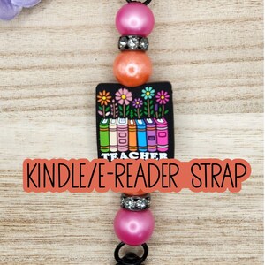 May include: A beaded strap featuring a black rectangular charm with a colorful illustration of books and flowers above the word TEACHER. The design includes pink and orange spherical beads, silver-toned rhinestone spacers, and black metal clasps.
