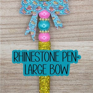 May include: A pen featuring a large blue bow with a pattern of ducks in swimming rings, topped with a yellow glitter bead. The barrel includes pink, turquoise, and gold beads, with a yellow glitter section and text reading RHINESTONE PEN-LARGE BOW.