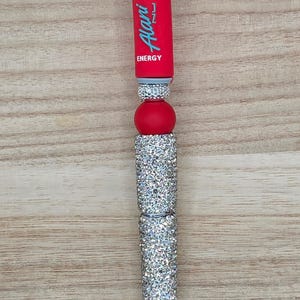K&ouml;nnte beinhalten: Ein individueller Stift mit rotem Schaft und dem hellblauen Schriftzug Alani ENERGY, verziert mit mehreren silbernen, strassbesetzten Perlen und einer glatten, roten Kugelperle. Der Stift verf&uuml;gt &uuml;ber eine silberfarbene Metallspitze.