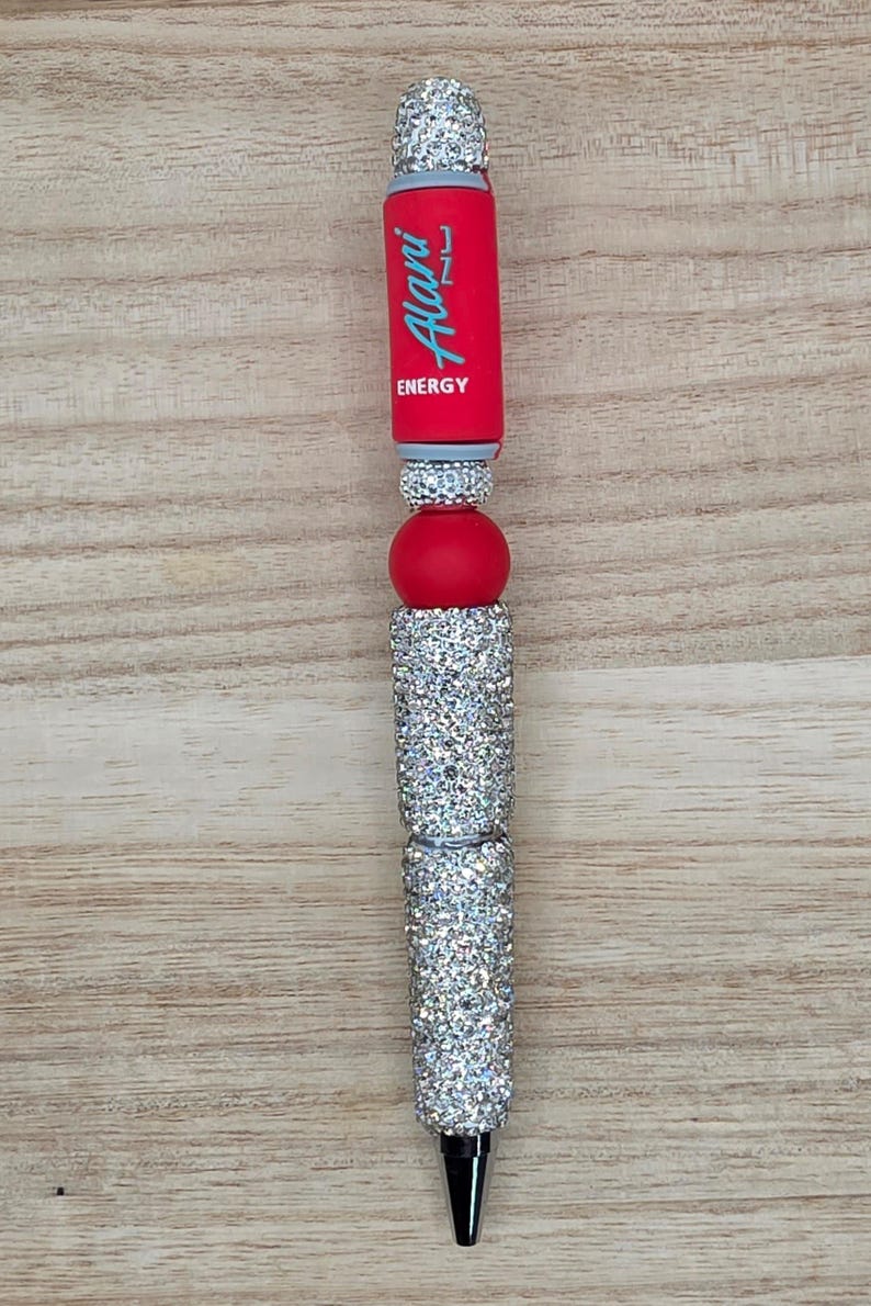 K&ouml;nnte beinhalten: Ein individueller Stift mit rotem Schaft und dem hellblauen Schriftzug Alani ENERGY, verziert mit mehreren silbernen, strassbesetzten Perlen und einer glatten, roten Kugelperle. Der Stift verf&uuml;gt &uuml;ber eine silberfarbene Metallspitze.