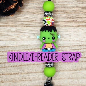 May include: A Kindle/E-reader strap featuring a central Frankenstein monster character bead, flanked by lime green round beads, silver-toned rhinestone spacers, and a multi-colored leopard print bead, all secured with black metal lobster claw clasps.