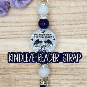 May include: A beaded Kindle or e-reader strap featuring a central circular charm with a glittery finish, a crescent moon, stars, mountain silhouettes, and the text "YOU KNOW WHAT THEY SAY ABOUT wingspan". It includes white and navy blue beads.