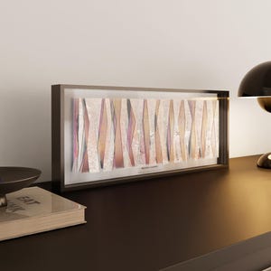 May include: A framed abstract artwork featuring vertical, irregular shapes in beige, pink, and brown hues. The piece is displayed on a dark surface, with a black desk lamp and a decorative bowl. The text "Aurora I London" is visible.
