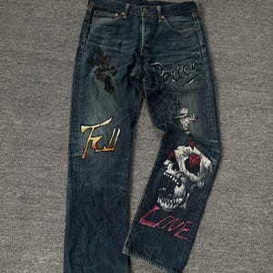 May include: Dark blue denim jeans with hand-painted designs. The designs include a skull with red eyes and diamonds, the words "Peace" and "Love", and a cross with the word "Evil" in yellow.
