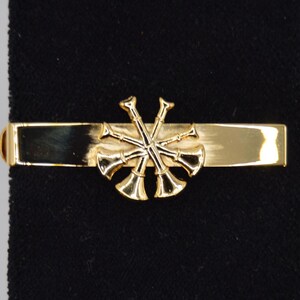 May include: A gold-toned tie clip featuring a cluster of crossed bugles. The clip has a rectangular bar with rounded edges and is set against a black background. This accessory is suitable for formal wear.