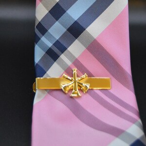 May include: Gold tie bar with a design of four crossed trumpets. The tie bar is on a pink and blue plaid tie.