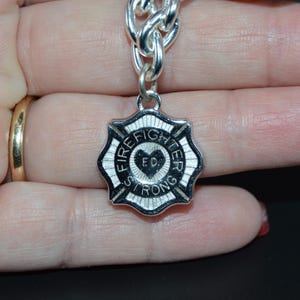 May include: A silver-colored charm with a firefighter's Maltese cross design. The charm has the words "FIRE FIGHTER STRONG" and "FD" with a heart in the center. The charm is attached to a silver chain.