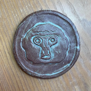 Könnte beinhalten: Ein runder, brauner Keramikuntersetzer mit einem erhabenen, stilisierten Affengesicht. Die Merkmale des Affen sind mit einer türkisfarbenen Glasur hervorgehoben. Ein einzigartiger dekorativer Artikel.