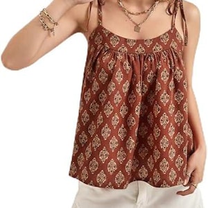 May include: A sleeveless, brown top with a repeating white floral pattern. The top has thin straps and a gathered neckline. The model is also wearing white shorts with frayed edges and gold jewelry.