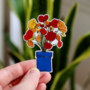 May include: A sticker featuring a bouquet of flowers in a blue pot. The flowers are red, orange, and yellow with green stems. The sticker is held in front of a blurred green background.
