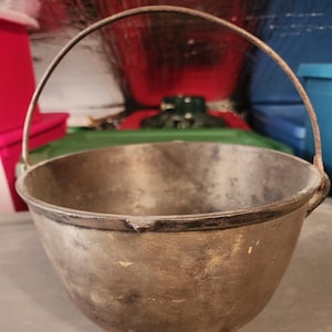 Puede incluir: Una olla de hierro fundido vintage con asa. La olla es de color marrón y tiene un aspecto desgastado y rústico.
