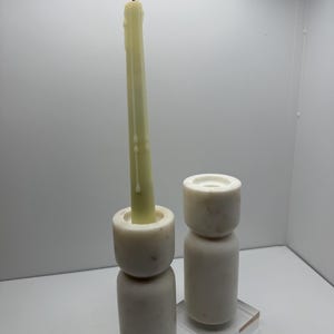 May include: Two white marble candle holders, one with a lit, off-white taper candle. The candle holders are cylindrical with a stacked design. The candle has a yellow flame. The second candle holder is empty and sits on a clear acrylic block.